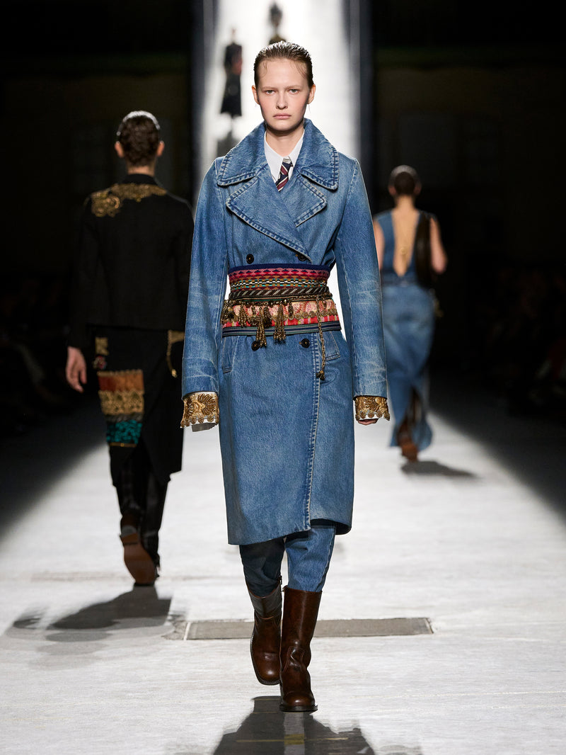 Model walking on a runway wearing a denim coat with a colorful belt.