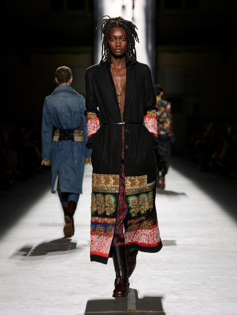 Model walking on a runway wearing a long, patterned coat with a dark background.