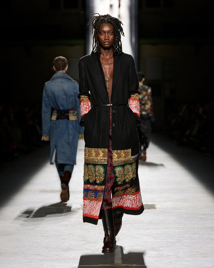 Model walking on a runway wearing a long, patterned coat with a dark background.