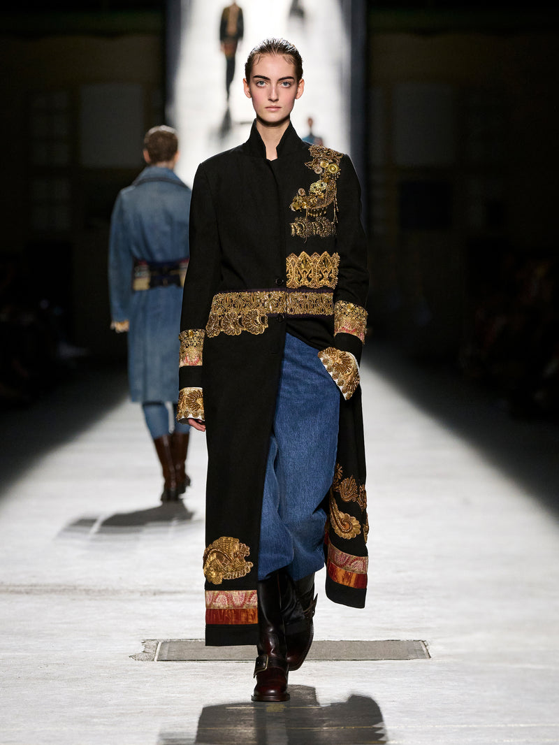 Model walking on a runway wearing a black coat with gold embroidery and blue pants.
