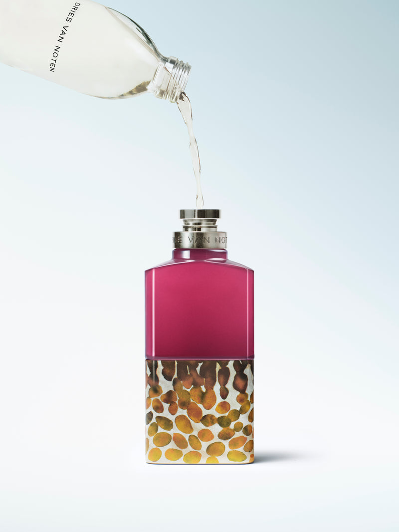 A perfume bottle with a magenta-pink upper section and a lower section filled with small gold, brown, and beige marbled beads, being refilled and shown on a light blue gradient background.