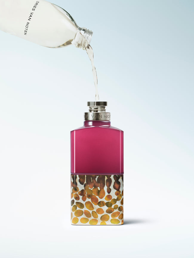 A perfume bottle with a magenta-pink upper section and a lower section filled with small gold, brown, and beige marbled beads, being refilled and shown on a light blue gradient background.