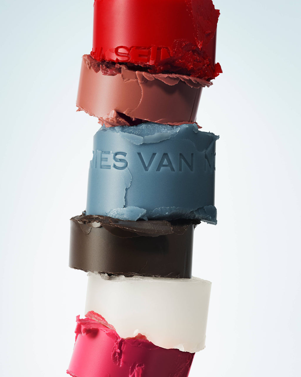 Stack of colorful lip balms with 'Dries Van Noten' branding on a light blue background