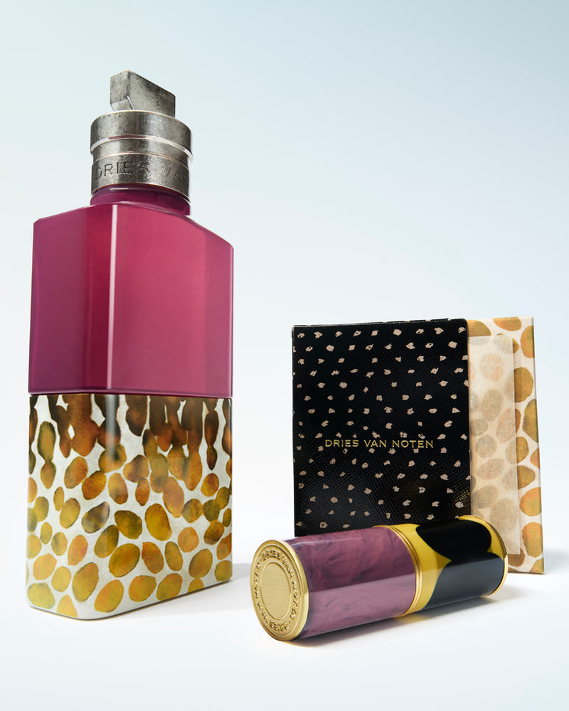A perfume bottle with a magenta upper section and a gold-and-white patterned lower section, shown alongside its black-and-gold patterned small box and a lip case cap on a light gray background
