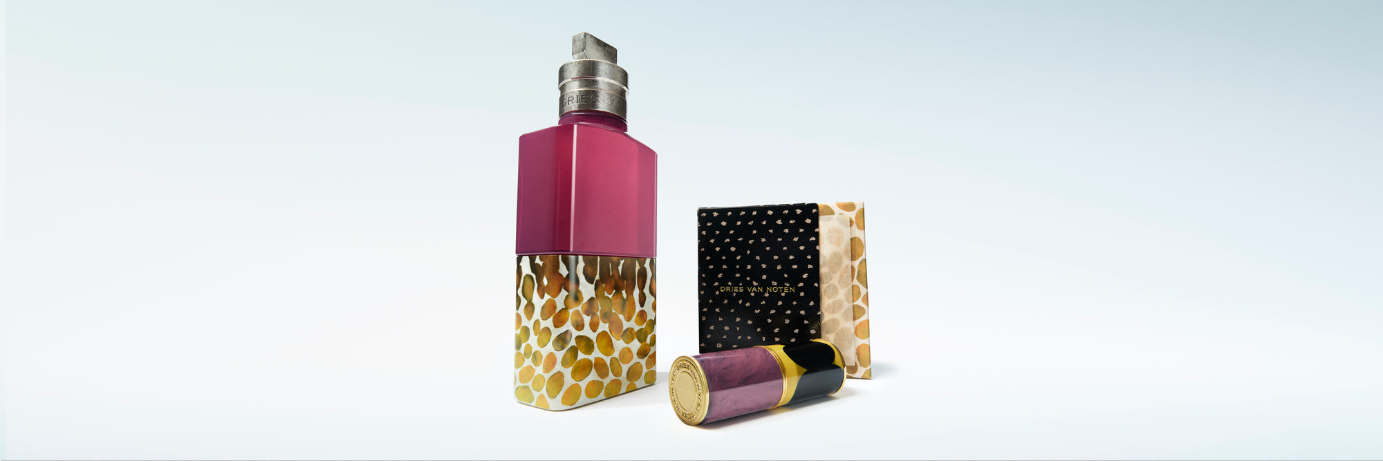 A fragrance bottle with a magenta upper section and a gold dot-print lower section, shown next to patterned box and a matching lipstick case on a light blue gradient background.