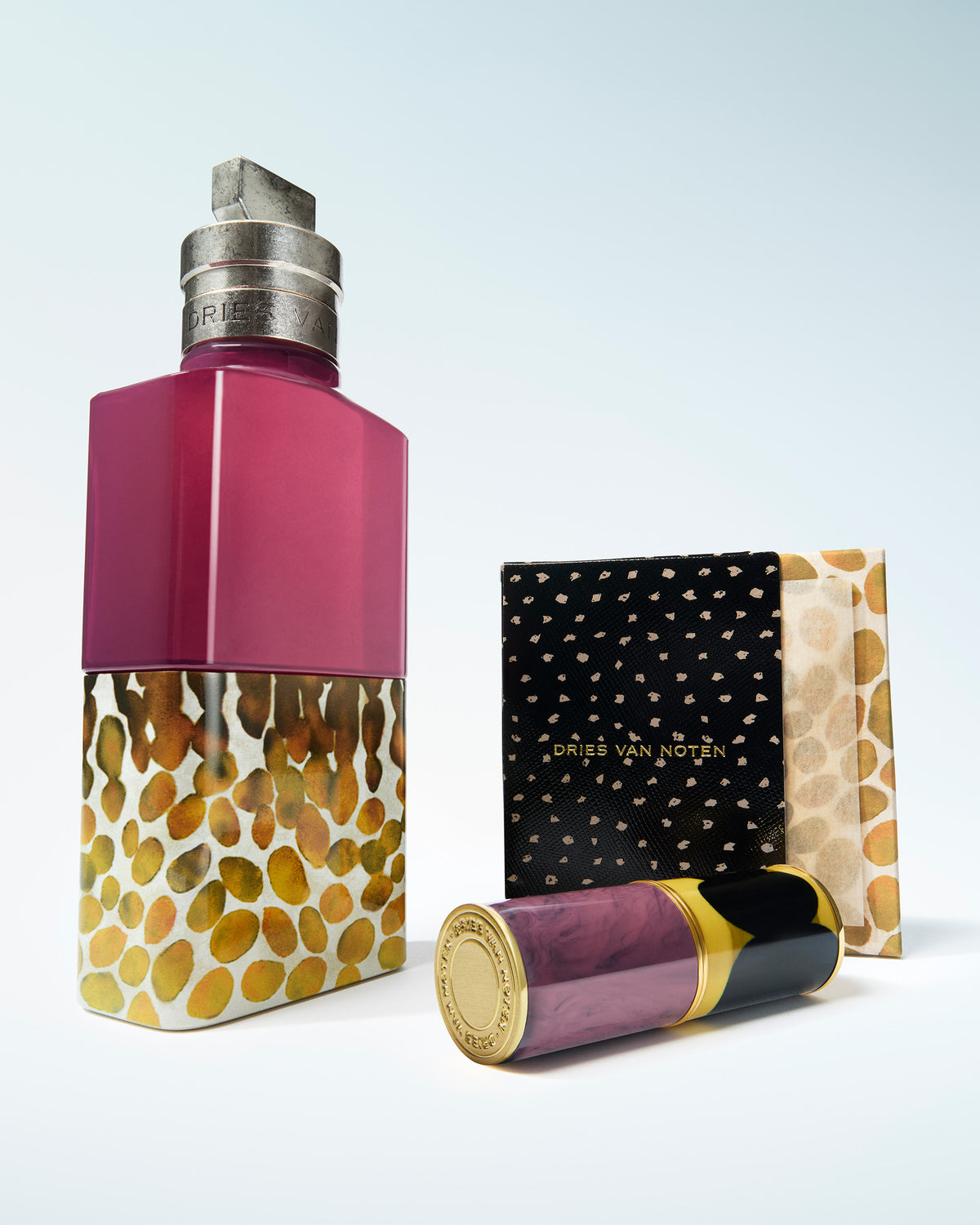 A Dries Van Noten set on the light blue-to-white gradient. background, featuring a magenta and gold fragrance bottle, a black carton box with gold dots and a matching purple and gold lip case.