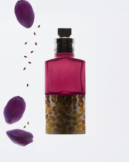 A fragrance bottle with a translucent magenta upper section and a patterned lower section featuring gold and brown organic spots on a white base shown against a clean white background with floating purple petals and small brown seeds on the left side.