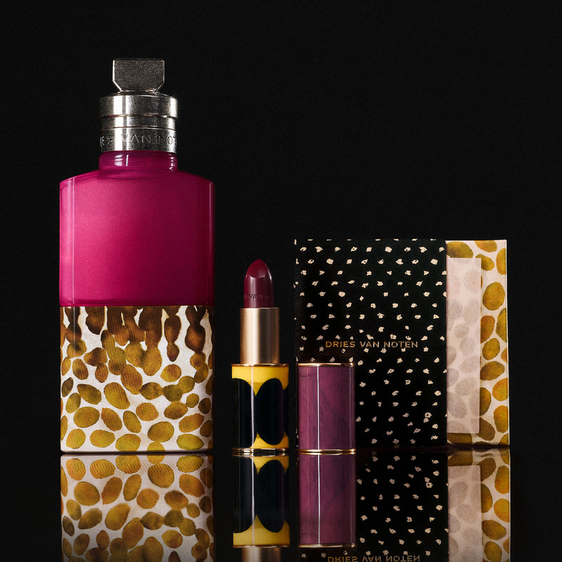 A cosmetics set featuring a large magenta and gold patterned fragrance bottle, a lipstick with a matching magenta cap, and a cupboard box on a reflective black surface.