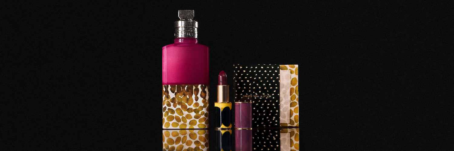 A cosmetics set featuring a large magenta and gold patterned fragrance bottle, a lipstick with a matching magenta cap, and a cupboard packaging boxes on a reflective black surface.