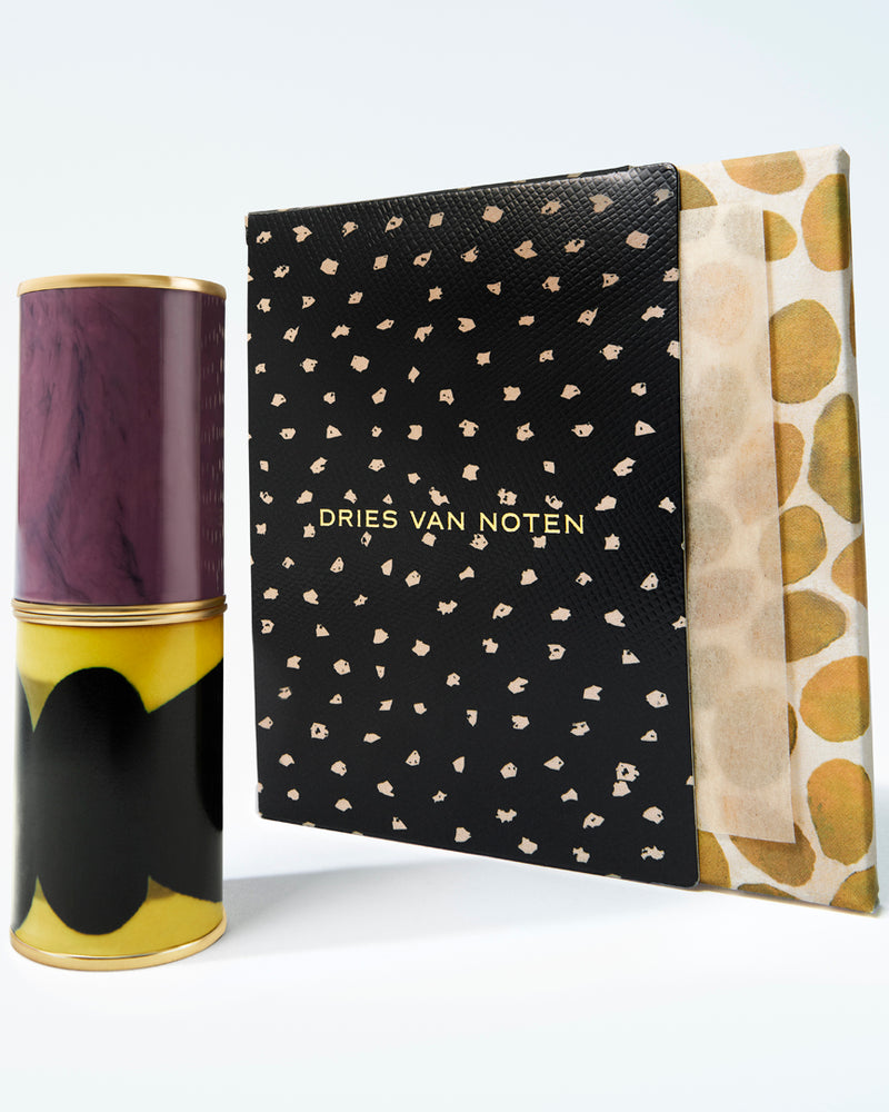A lipstick case with a purple and yellow marbled print, standing next to its black patterned box with gold lettering and a dotted-print side panel.