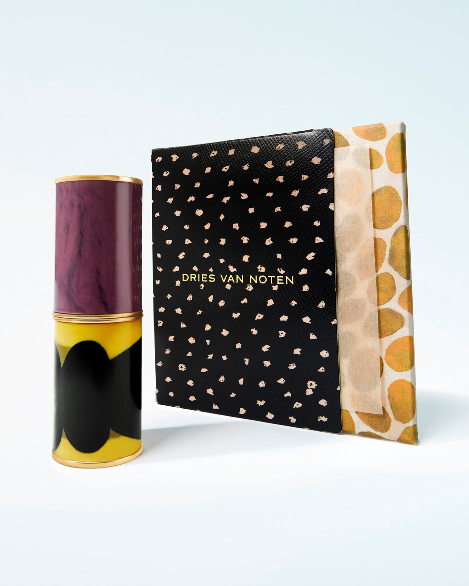 Dries Van Noten cosmetics set featuring a lipstick case, next to a black patterned carton box with black and gold studs on a light blue gradient background.