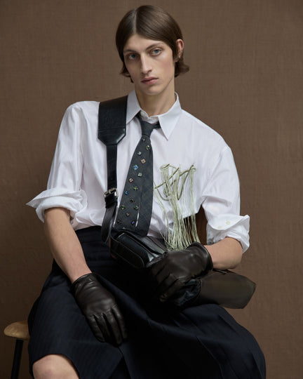Seated model wearing a white shirt with green fringe embroidery, an embellished gray tie, dark tailored shorts, leather gloves, and a black crossbody bag.