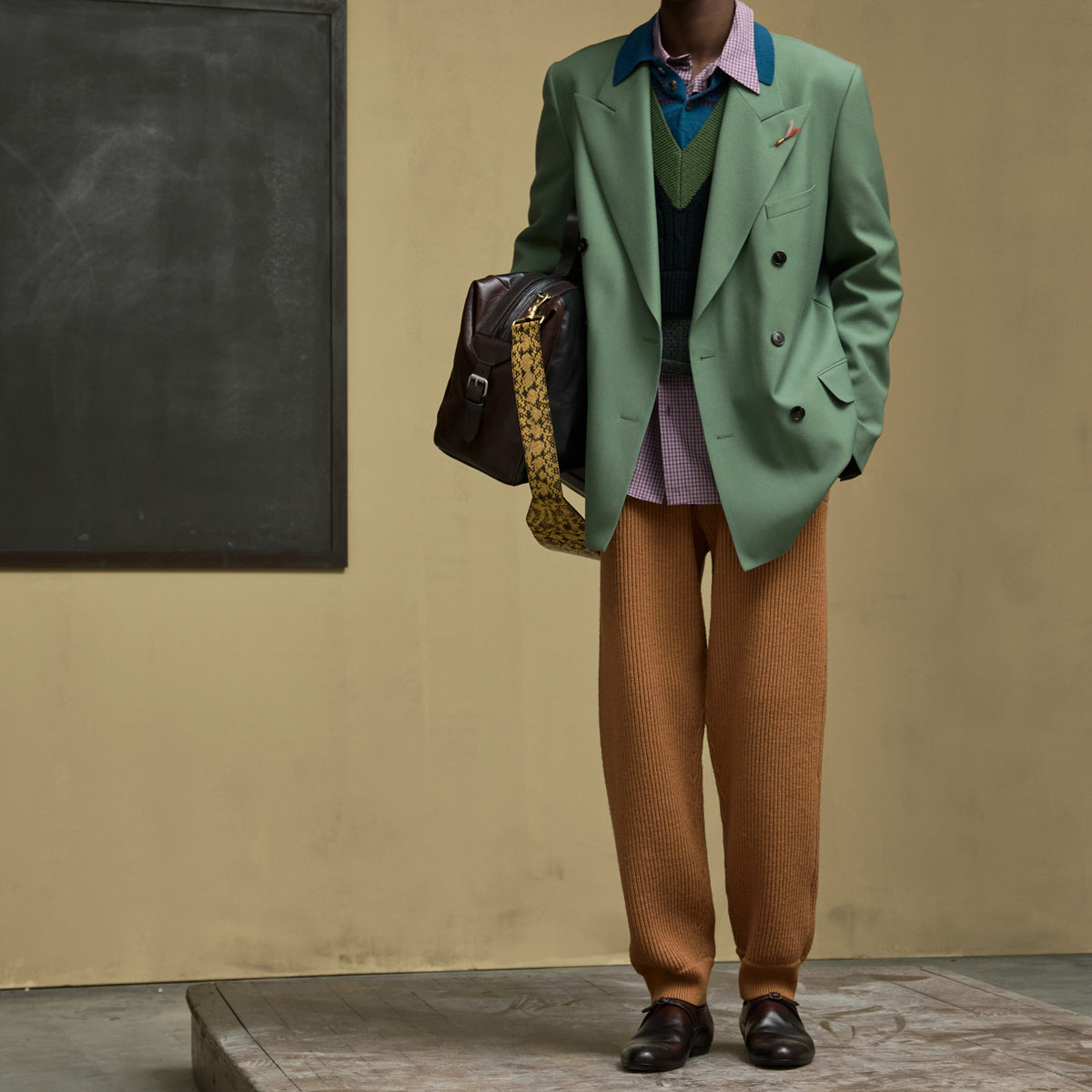Model wearing a sage green double-breasted blazer, rust-brown textured trousers, a green and navy sweater vest, a pink patterned shirt, and dark brown leather shoes, holding a dark brown shoulder bag with a patterned strap, standing against a beige wall with a chalkboard.