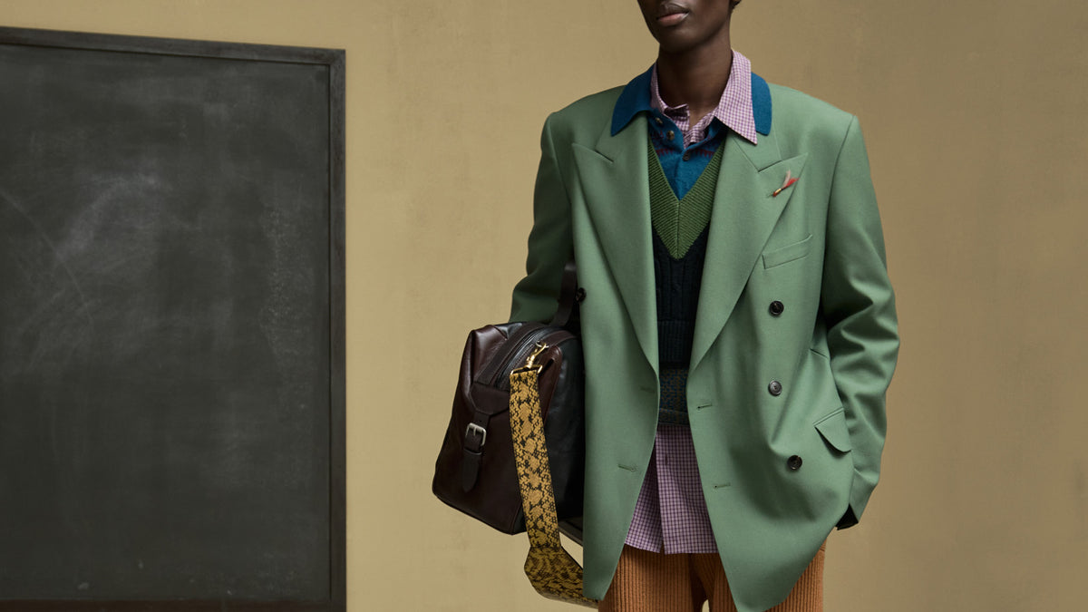 Model wearing a sage green double-breasted blazer over a green knit sweater vest, a pink patterned shirt, and brown trousers, holding a dark brown leather shoulder bag with a patterned strap against a beige wall and blackboard background.
