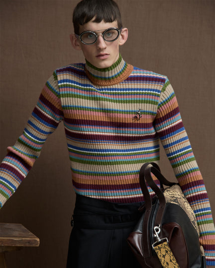 Model wearing oval glasses, a multicolored striped turtleneck sweater, and a leather bag with snakeskin details.