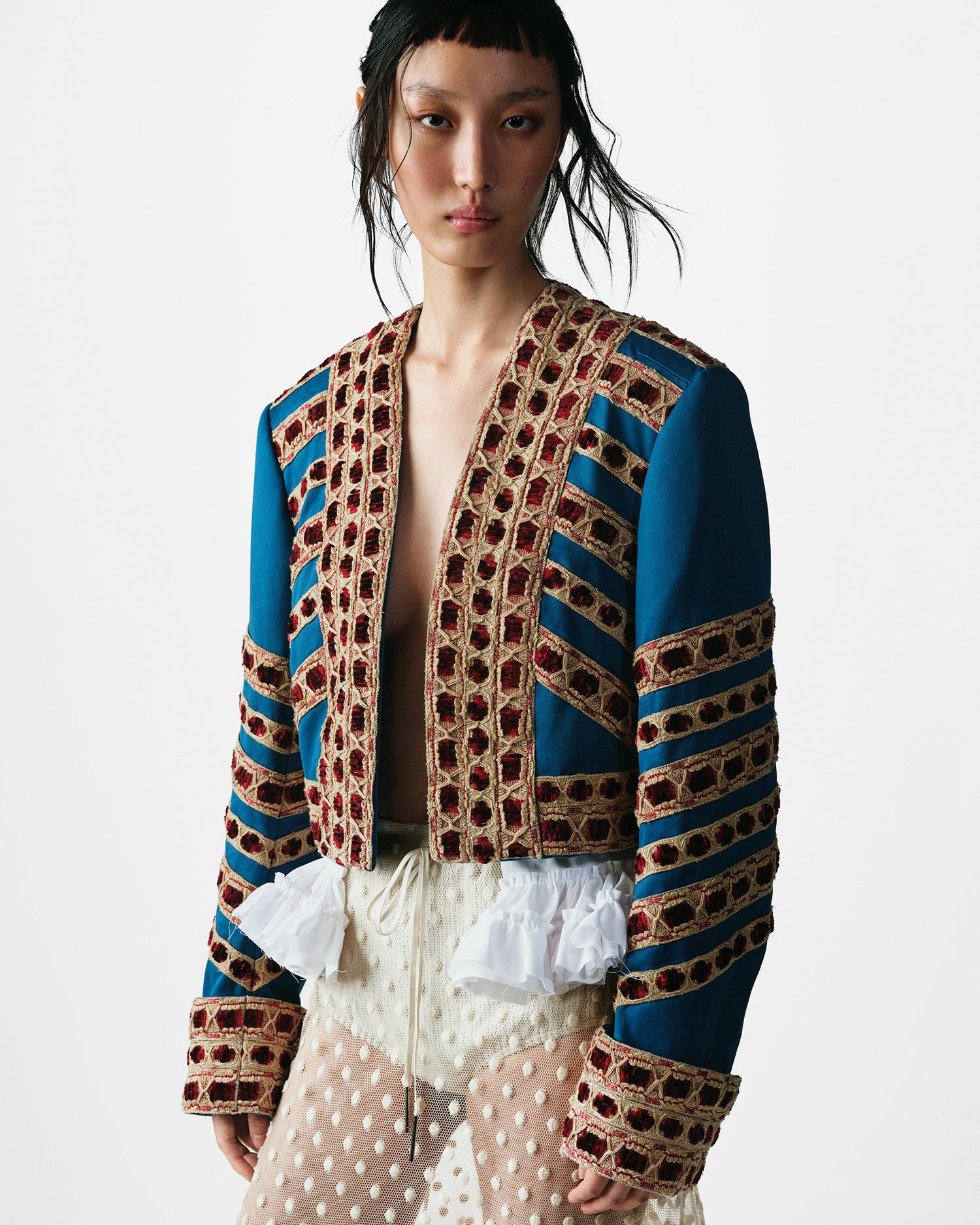 Model wearing a structured blue jacket and red embroidered detailing, styled with a sheer textured skirt 