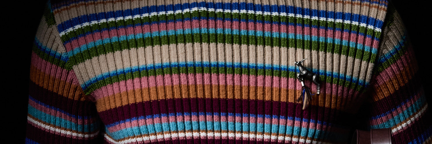 A close-up of a ribbed knit sweater featuring horizontal multicolor stripes i with a small pin on the chest.
