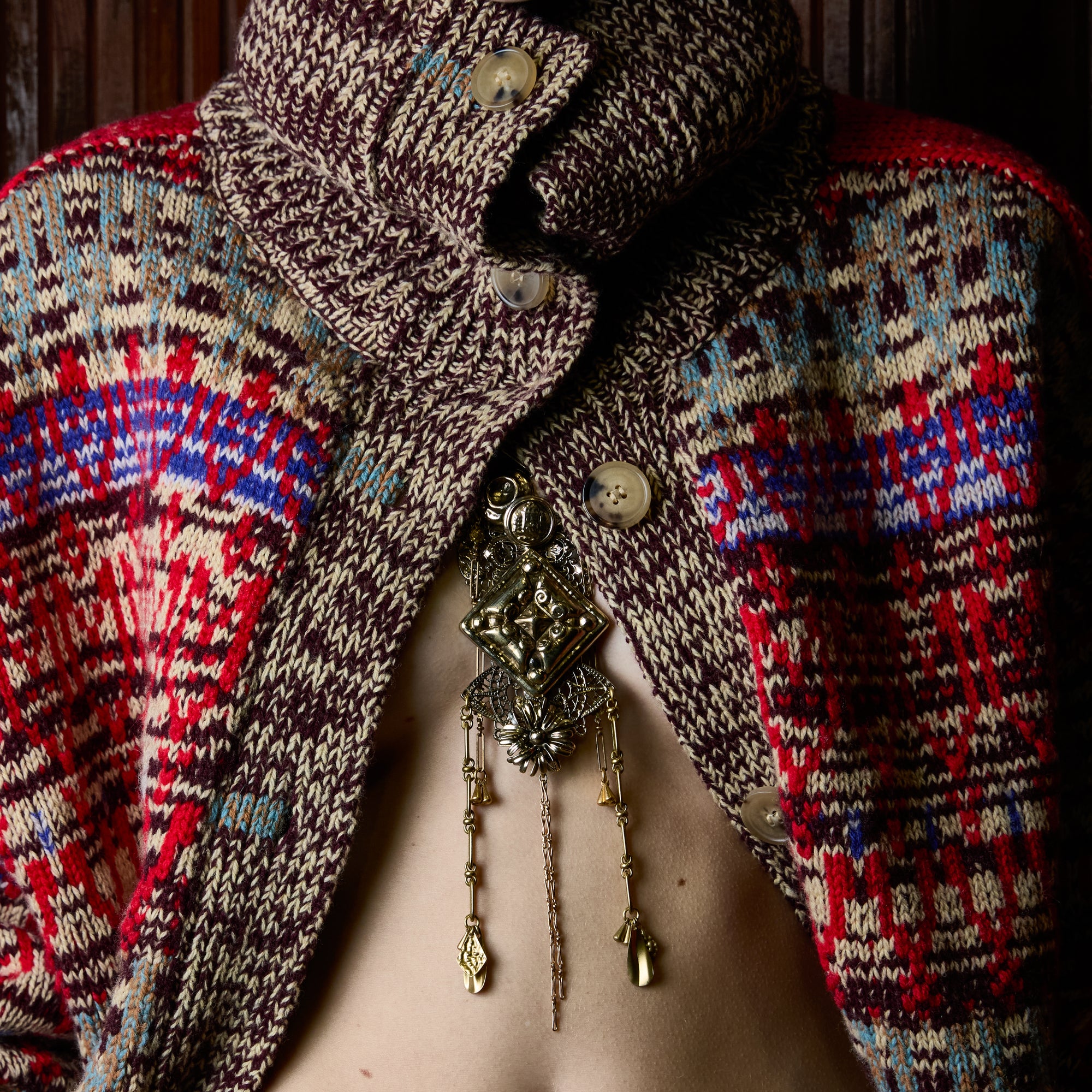 A close-up of a person wearing a heavily textured, multicolor knit cardigan with a high folded collar and ornate gold-toned jewelry draped over the chest.