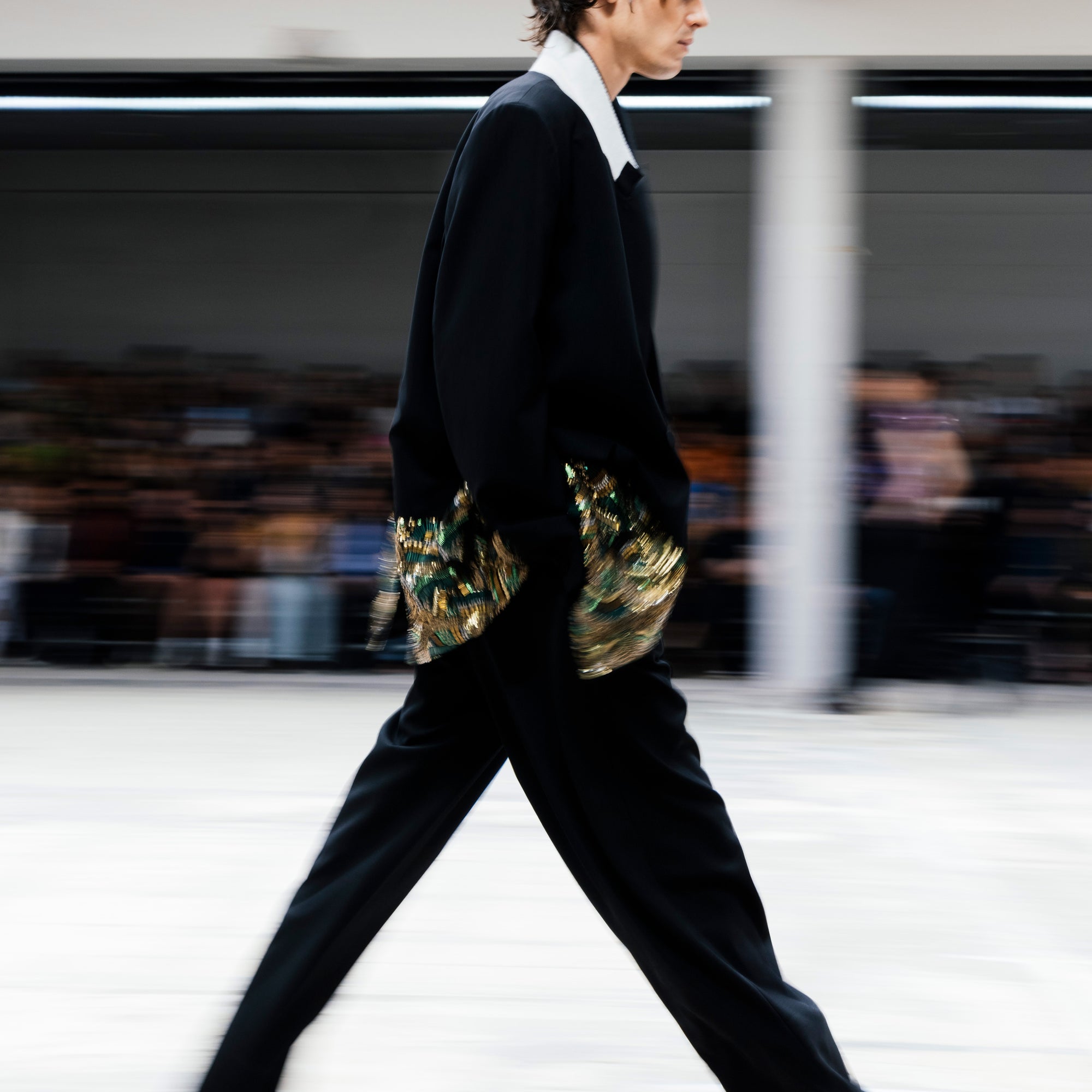 Model walking on a runway, wearing  a black suit with gold and green embroidery accents with a blurred audience in the background