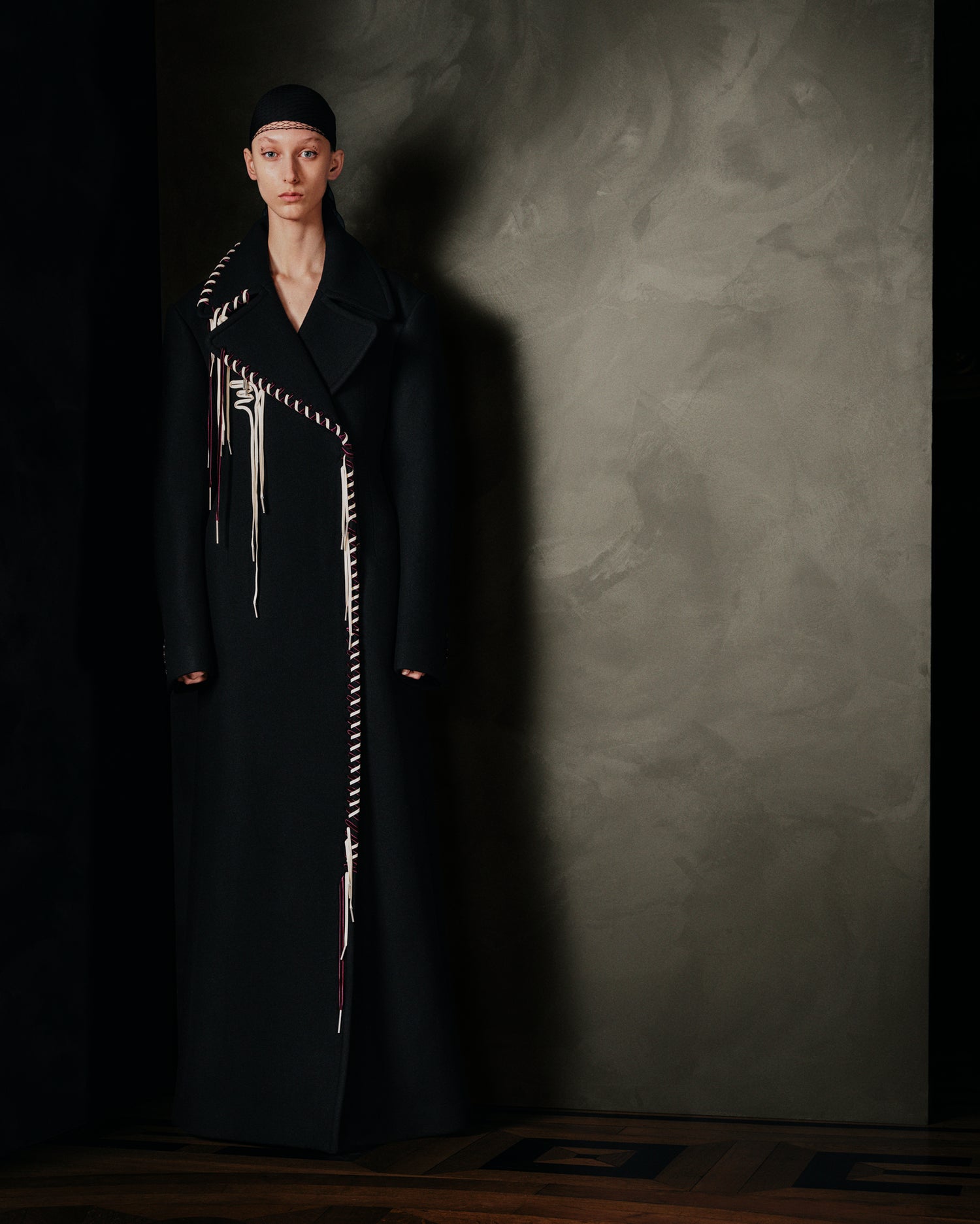Person wearing a long black coat with unique patterns against a textured gray wall.