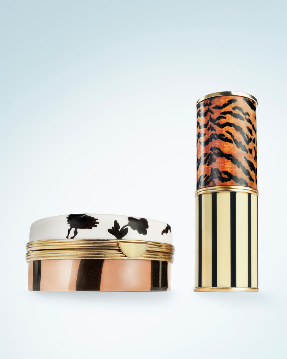 A luxury cosmetic container set featuring a round compact with a white lid decorated with black animal silhouettes and a metallic rose gold base, alongside a matching cylindrical tube with an orange and black tiger stripe pattern on the upper half and vertical black stripes on the metallic rose gold lower half, set against a light blue gradient background.