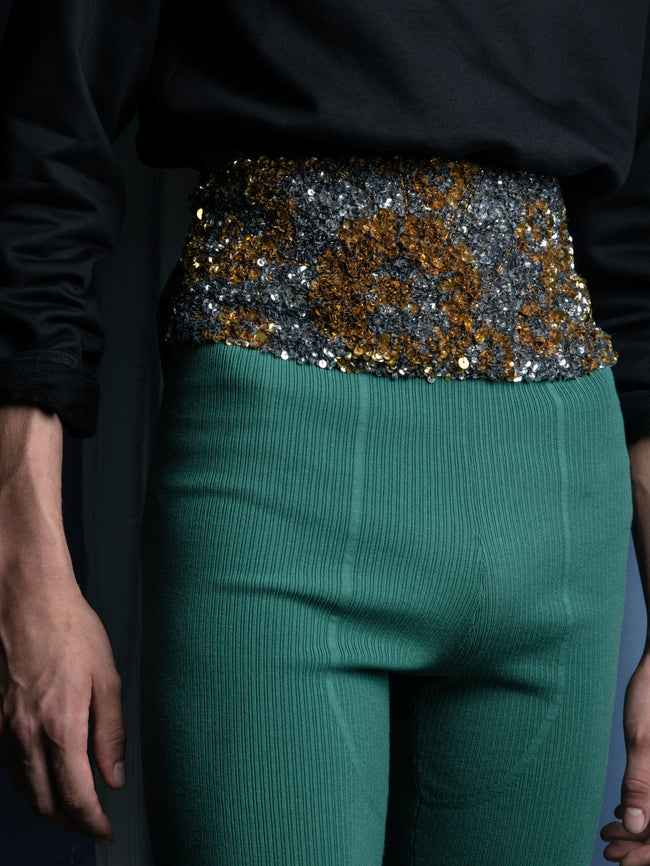 Person wearing a black long-sleeve top with a sequined waistband and green pants against a dark background.