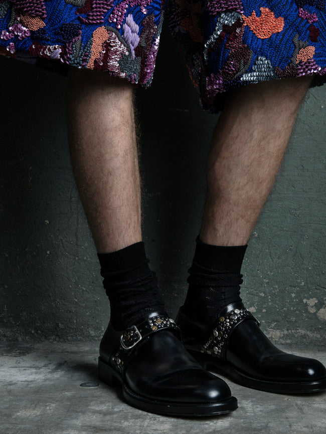 Person wearing black leather boots with embellishments, black socks, and a colorful sequined skirt on a dark background.