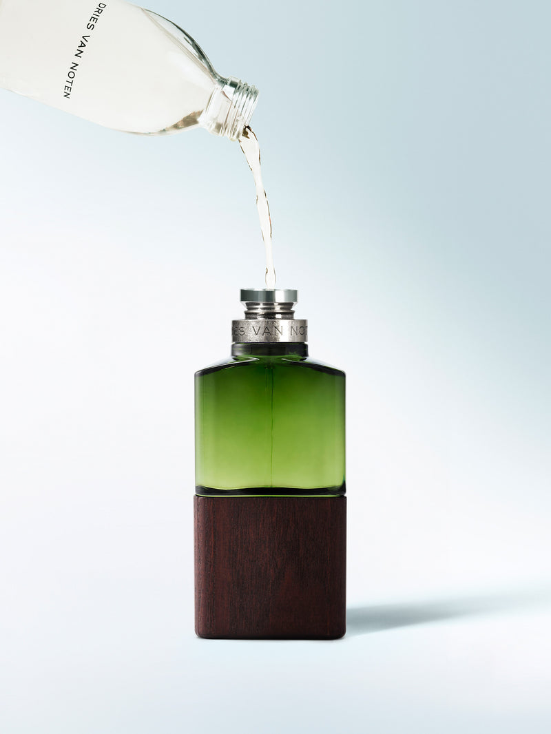 Green bottle with wooden base being filled with liquid from a white container on a light blue background