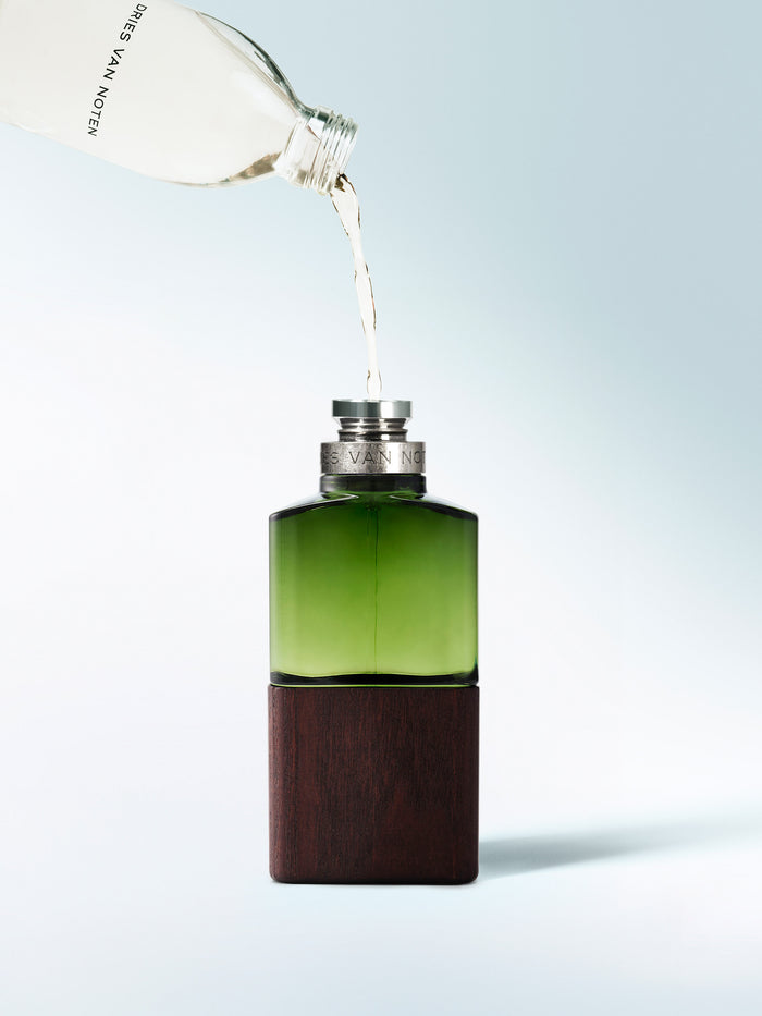 Green bottle with wooden base being filled with liquid from a white container on a light blue background