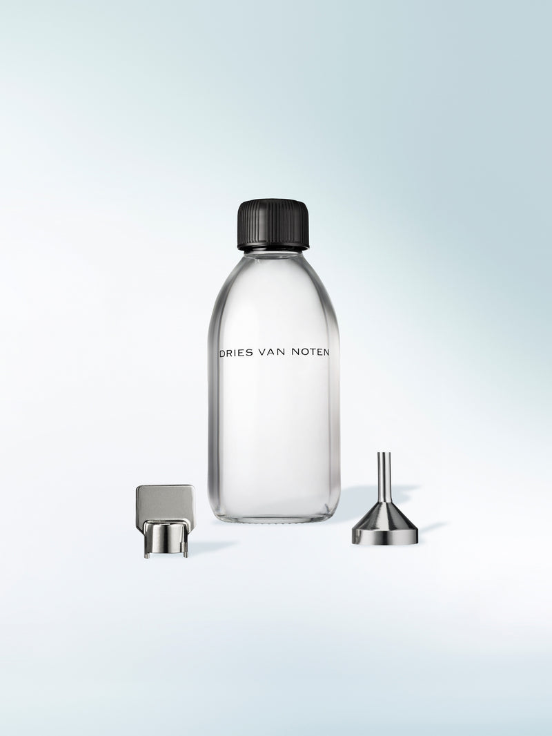 A clear glass labeled perfume refill bottle with a black screw cap, shown on a light blue gradient background with a metal pour spout and funnel accessory placed beside it.