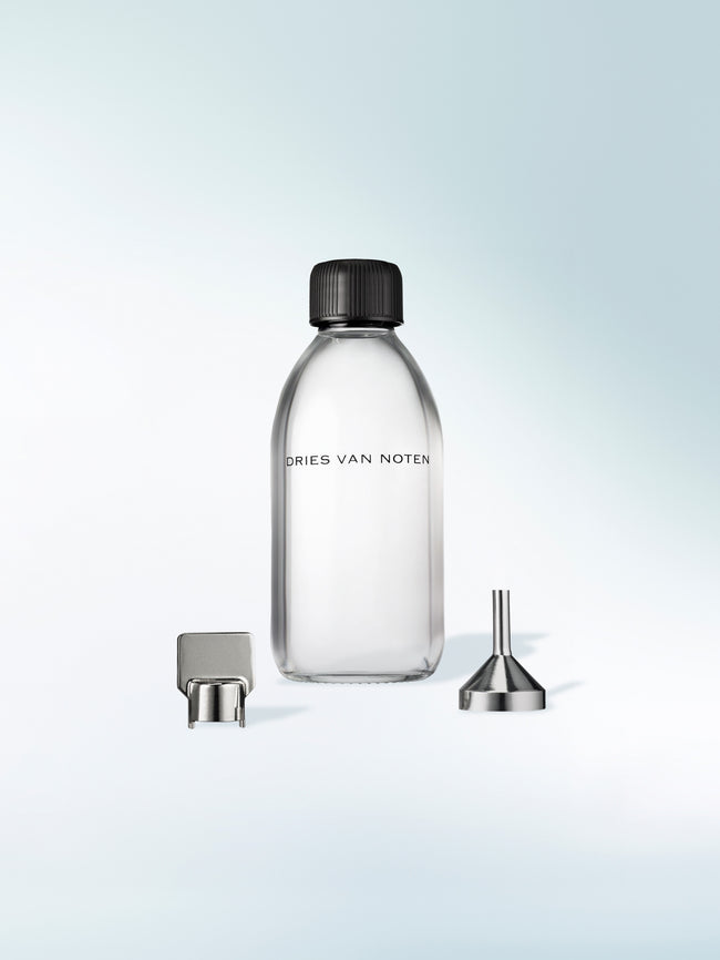 A clear glass labeled perfume refill bottle with a black screw cap, shown on a light blue gradient background with a metal pour spout and funnel accessory placed beside it.