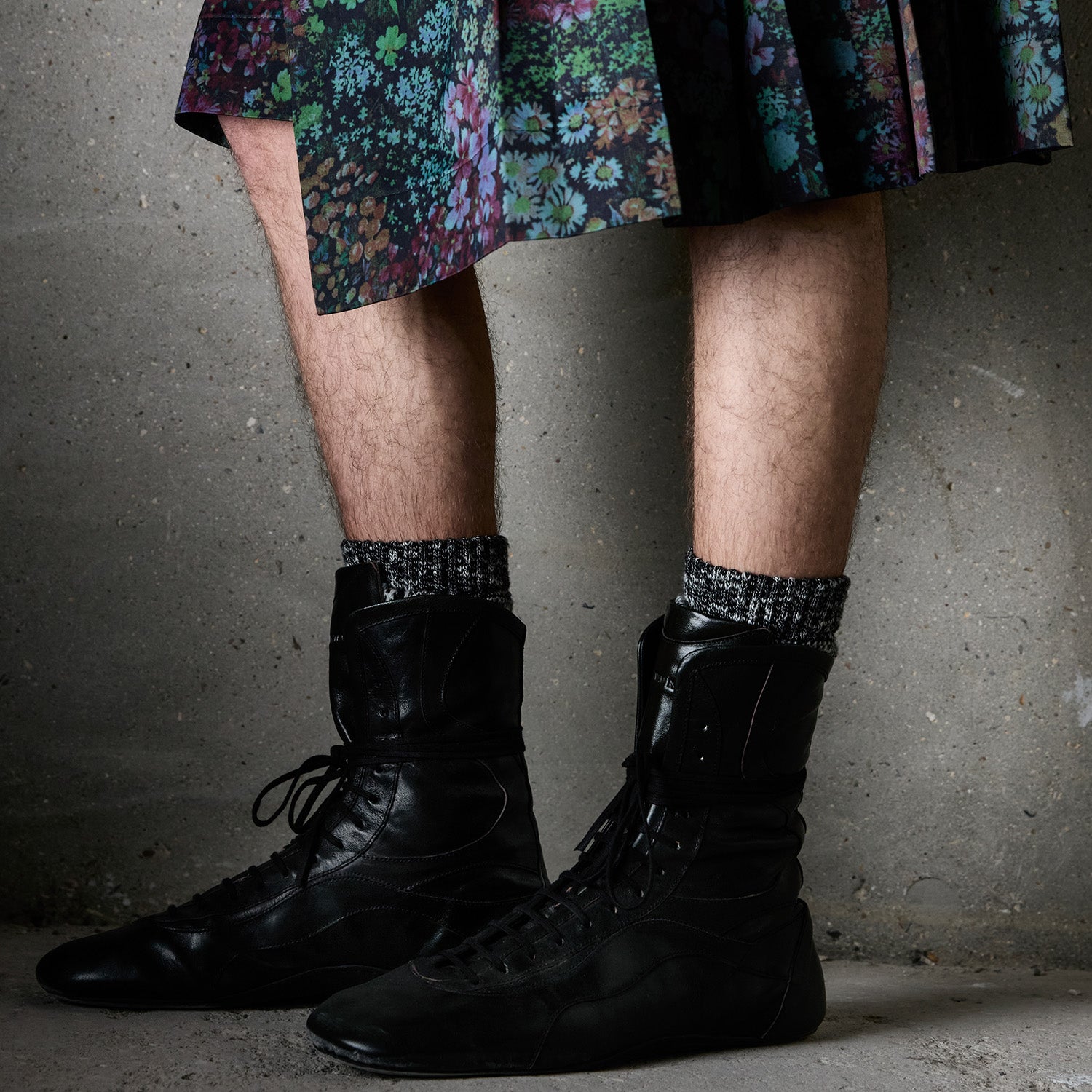 A close-up of a person's legs wearing black lace-up ankle shoes, patterned knit socks and a dark pleated skirt with a colorful floral print, standing against a textured concrete wall. 