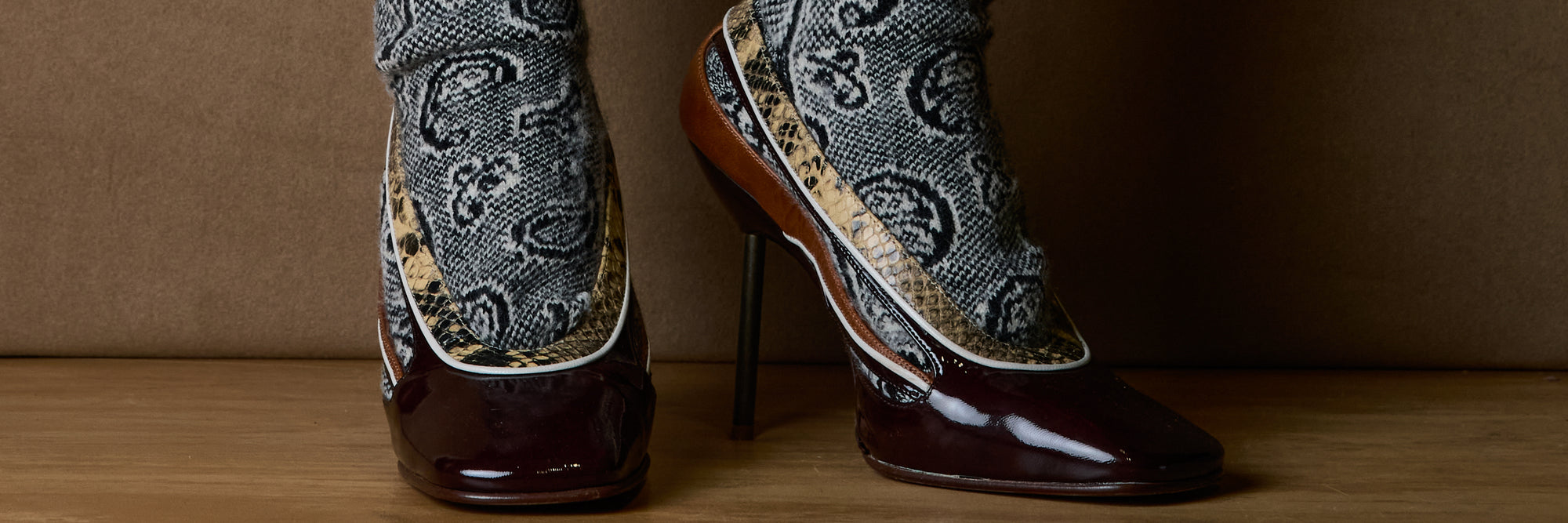 A pair of high-heeled boots with glossy dark brown patent leather toes and heels, decorated with gray and black paisley-patterned socks, shown on a wooden surface against a brown background.