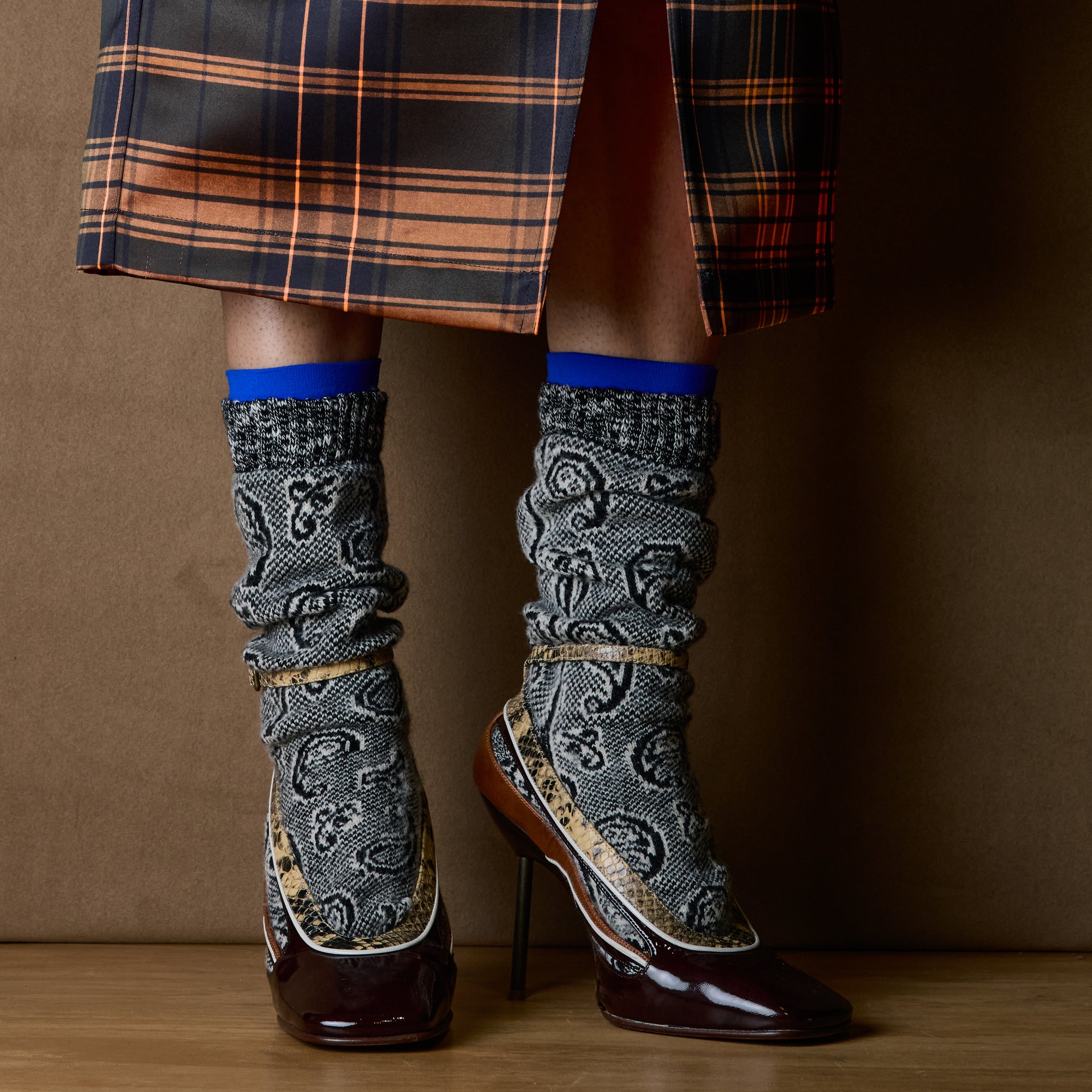 A person wearing knee-high patterned socks with a glossy dark brown pointed-toe heels, shown from the mid-thigh down against a brown studio backdrop.
