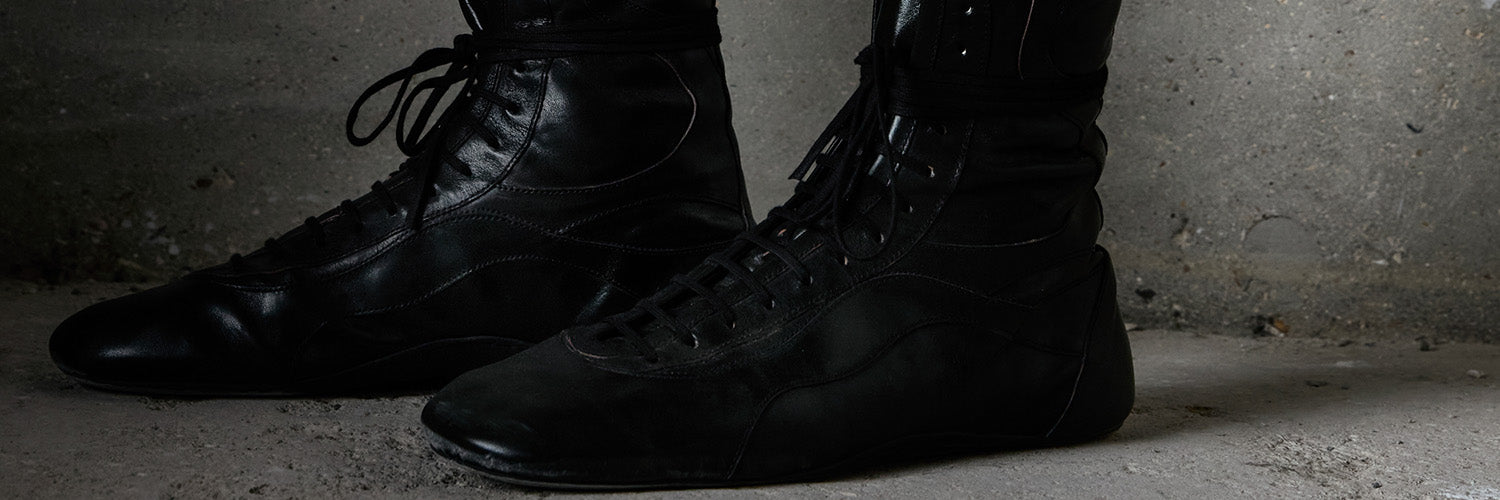 A pair of black leather high-top racing boots with laces, shown from the side against a concrete background.