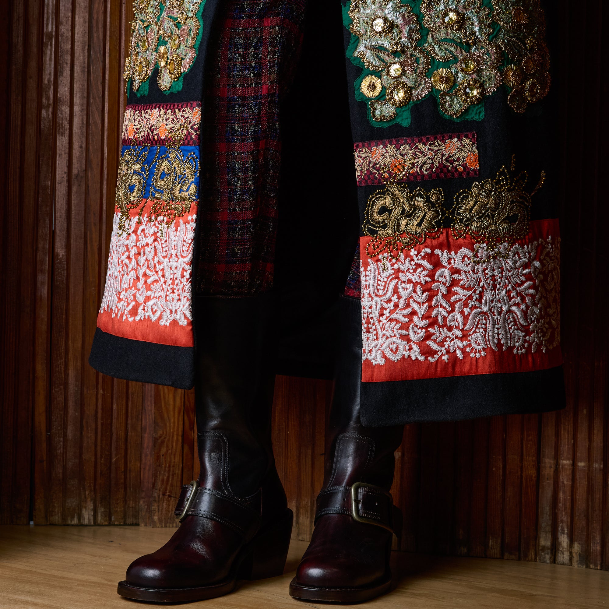 A close-up of a person's lower legs wearing dark brown leather buckle-strap boots and a highly detailed, embroidered black coat with red, green, and gold patterns, standing against a dark wood-paneled wall.