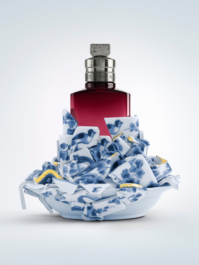 Bottle with a red label on a decorative blue and white ceramic base against a light gray background - Dries Van Noten