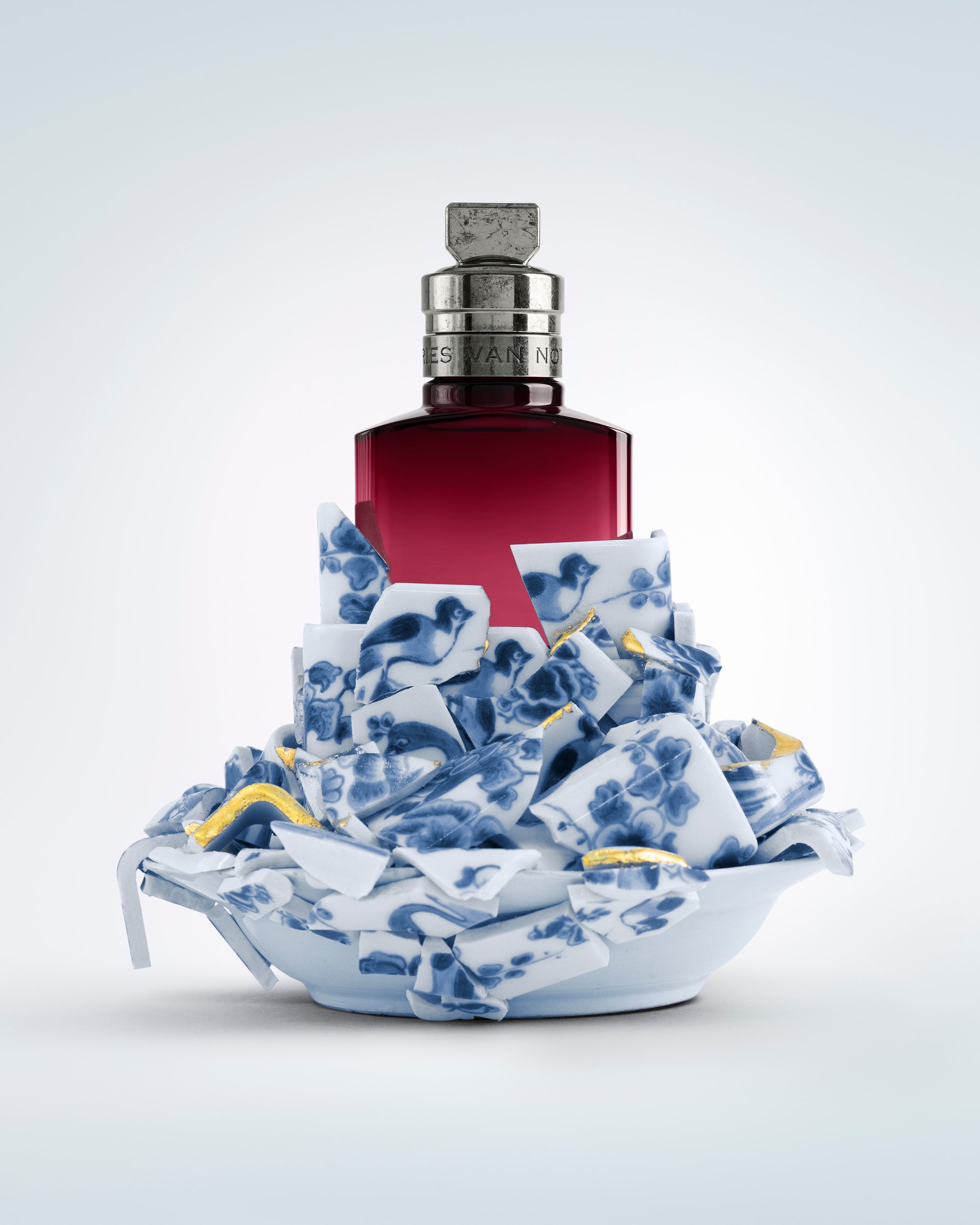 Bottle with a red label on a decorative blue and white ceramic base against a light gray background - Dries Van Noten