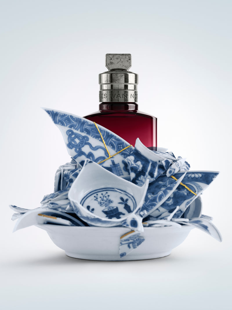 Broken blue and white ceramic pieces with a red bottle on a light gray background - Dries Van Noten