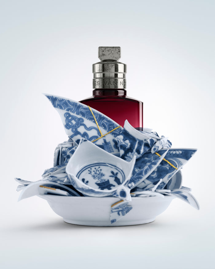 Broken blue and white ceramic pieces with a red bottle on a light gray background - Dries Van Noten