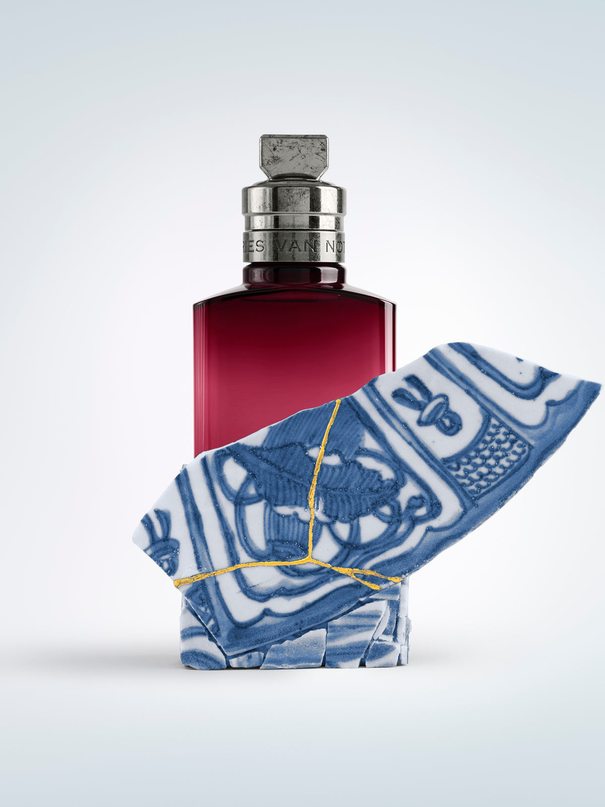 Red perfume bottle with a blue and white patterned cloth on a light gray background - Dries Van Noten