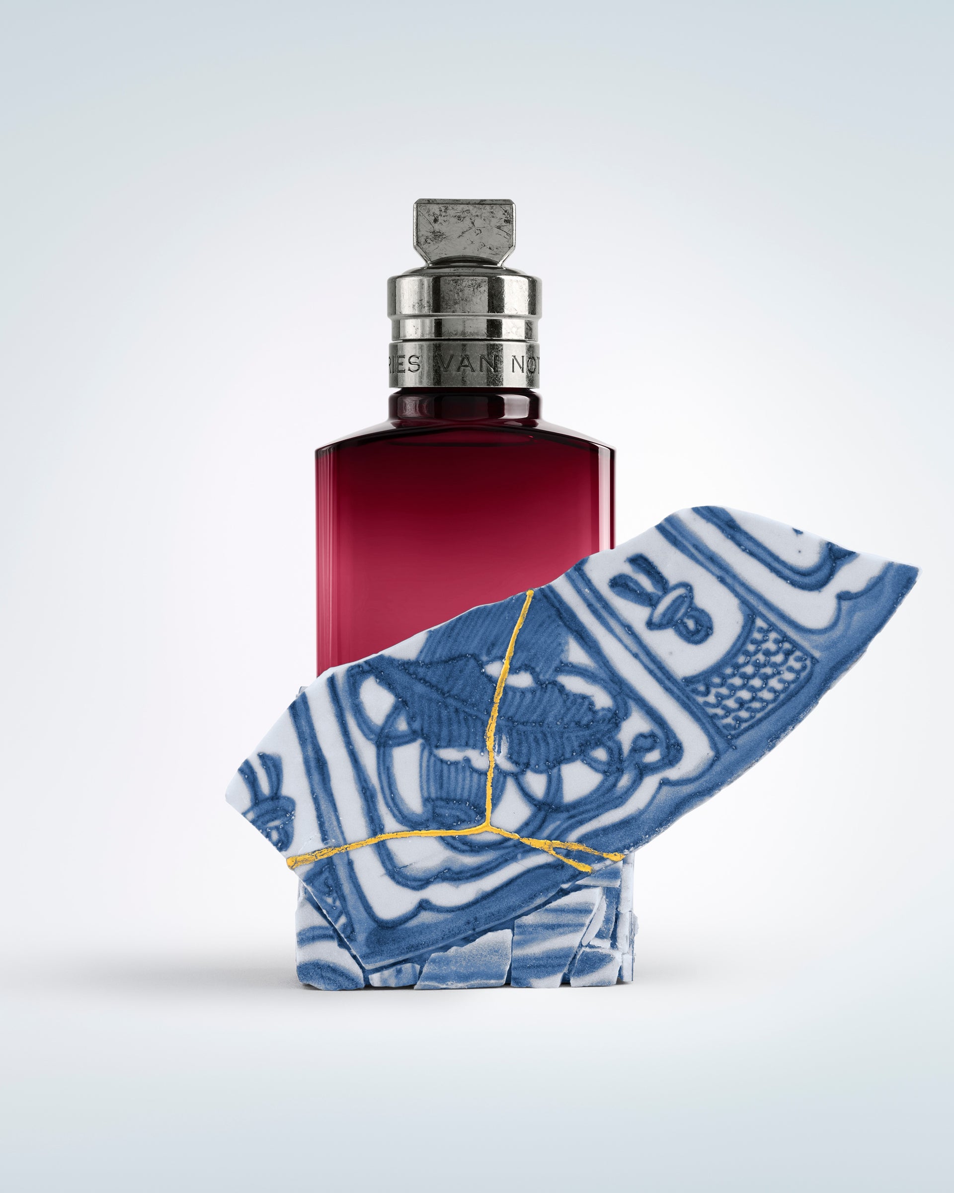 Red perfume bottle with a blue and white patterned cloth on a light gray background - Dries Van Noten