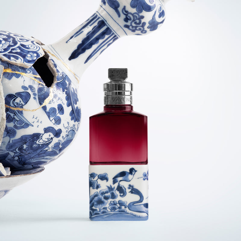 Red perfume bottle with blue and white decorative elements on a light gray background - Dries Van Noten