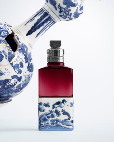 Perfume bottle with blue and white decorative design next to a ceramic teapot on a white background - Dries Van Noten