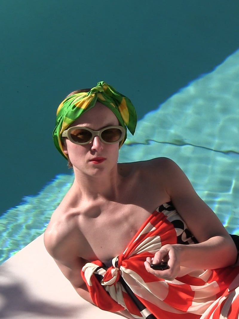 Person wearing oval sunglasses and a green patterned headscarf reclining beside a swimming pool an a red patterned pareo 
