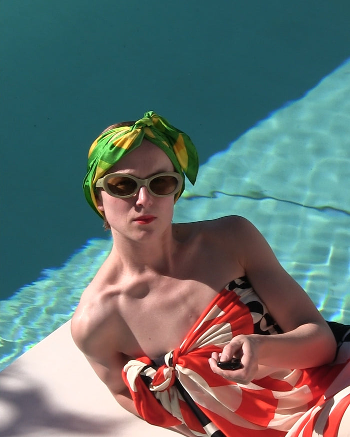 Person wearing oval sunglasses and a green patterned headscarf reclining beside a swimming pool an a red patterned pareo 