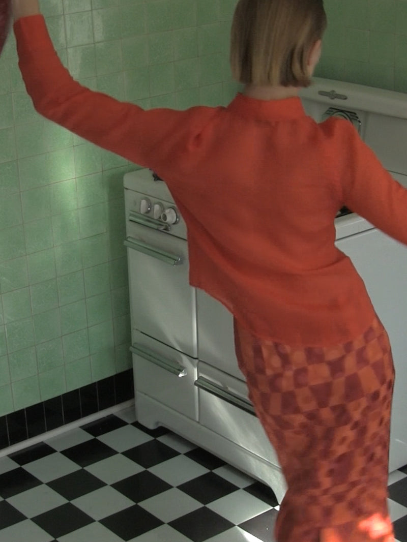 Person in orange outfit standing in a kitchen with green tiled walls and black and white checkered floor.