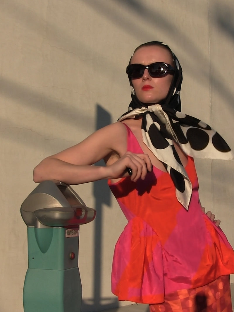Woman in a colorful set  and sunglasses interacting with a parking meter against a plain wall.