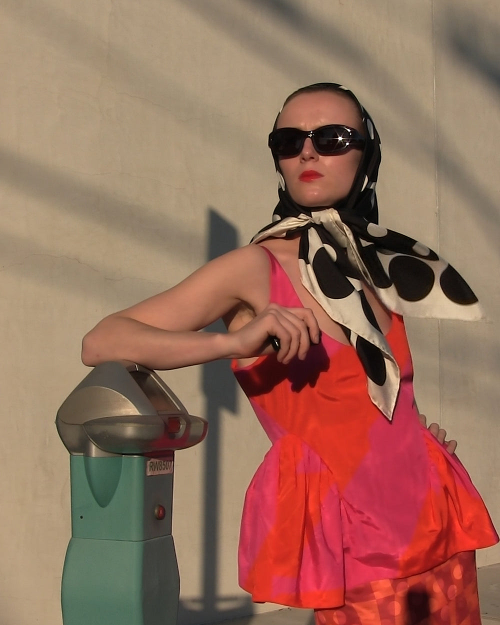 Woman in a colorful set  and sunglasses interacting with a parking meter against a plain wall.