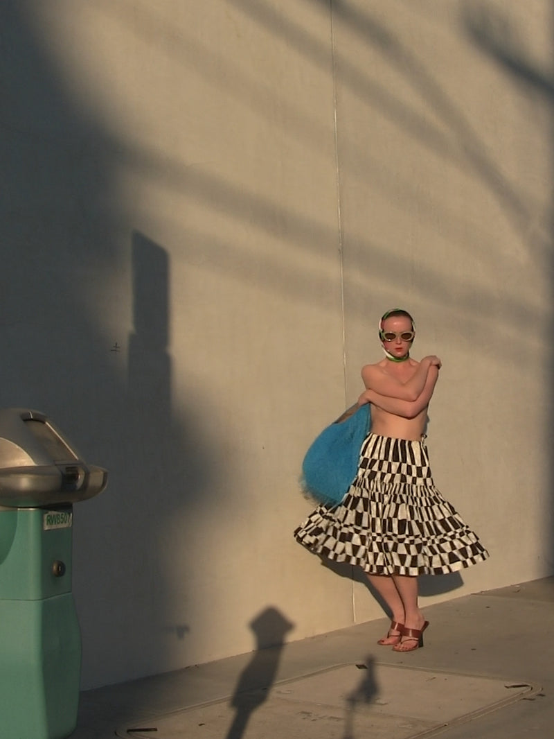 Woman in a checkered skirt and sunglasses standing against a wall with a shadow. 